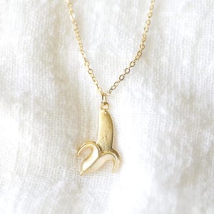 May include: A gold-coloured necklace featuring a pendant in the shape of a peeled banana. The banana pendant hangs from a fine chain. The necklace is set against a white, textured backdrop.