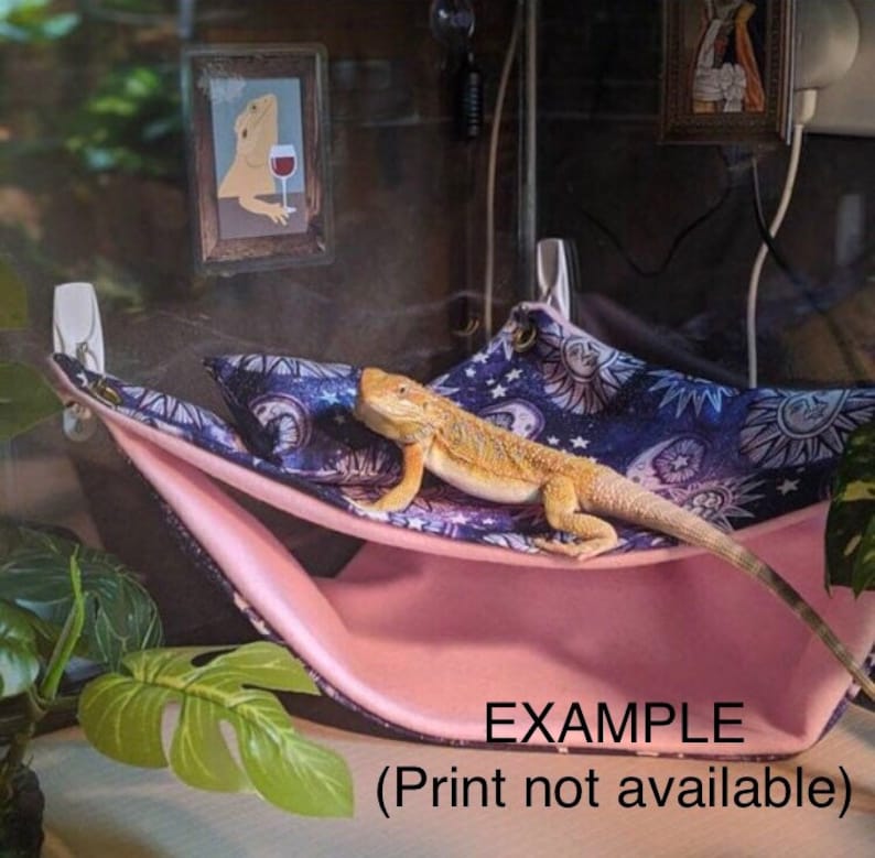 May include: A pink and blue hammock with a celestial print and a bearded dragon lizard resting on it. The hammock is attached to the side of a glass terrarium. The text "EXAMPLE (Print not available)" is printed on the hammock.