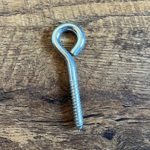 May include: A single silver-colored eye bolt with a threaded screw end. The eye is a closed loop, and the screw portion has a pointed tip. The eye bolt is set against a brown wood grain background.