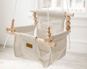 May include: A white and gray striped fabric baby swing with wooden dowels and wooden beads. The swing is hanging from white ropes and has a "Sweet Swinging" label on the fabric.