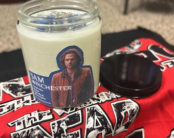 Op de afbeelding: Een glazen potkaars met een zwarte deksel, met het label "Sam Winchester" en een afbeelding van een man. De kaars staat op een rood en zwart bedrukte stof.