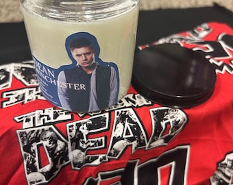 May include: A clear glass jar candle with a black lid, featuring a label with a man's image and the text "DEAN WINCHESTER". The candle sits on a red and black fabric with the words "THE DEAD" in a stylized font.