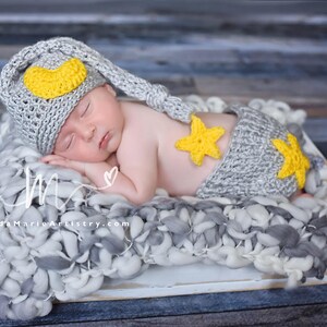 Puede incluir: Un bebé recién nacido con un gorro de dormir de crochet gris con una luna creciente amarilla y un pañal de crochet gris a juego con estrellas amarillas. El bebé está durmiendo en una manta peluda gris y blanca.