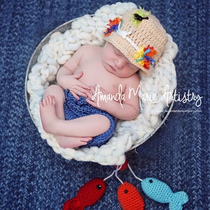 May include: A newborn baby wearing a crocheted fishing hat with colourful accents, sleeping in a white bucket with a blue knitted nappy. Three crocheted fish hang below the bucket on a blue background.