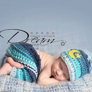 May include: A sleeping baby wearing a blue and white crocheted hat with a yellow crescent moon and star applique. The baby is wearing a blue and white crocheted swaddle. The baby is lying on a blue and white blanket. The image includes the text "Once Upon A Dream Photography".