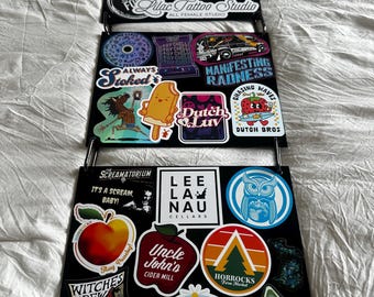 May include: A black, three-tiered organizer with a gold chain handle, covered in colorful stickers. The stickers feature various designs, including logos, illustrations, and text. The organizer is displayed on a white, textured surface.