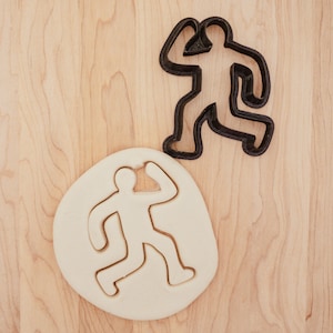 May include: A white cookie cutter in the shape of a person, with a black cookie cutter in the shape of a person on a wooden surface.