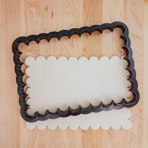 May include: A black, scalloped-edge cookie cutter sits on a piece of pale dough, with a second piece of dough beneath. The cutter is rectangular with a decorative edge. The background is a light wood surface.