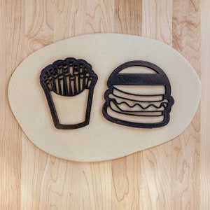 May include: A close-up of two black cookie cutters on a light brown surface. The cookie cutters are shaped like a hamburger and a container of french fries.