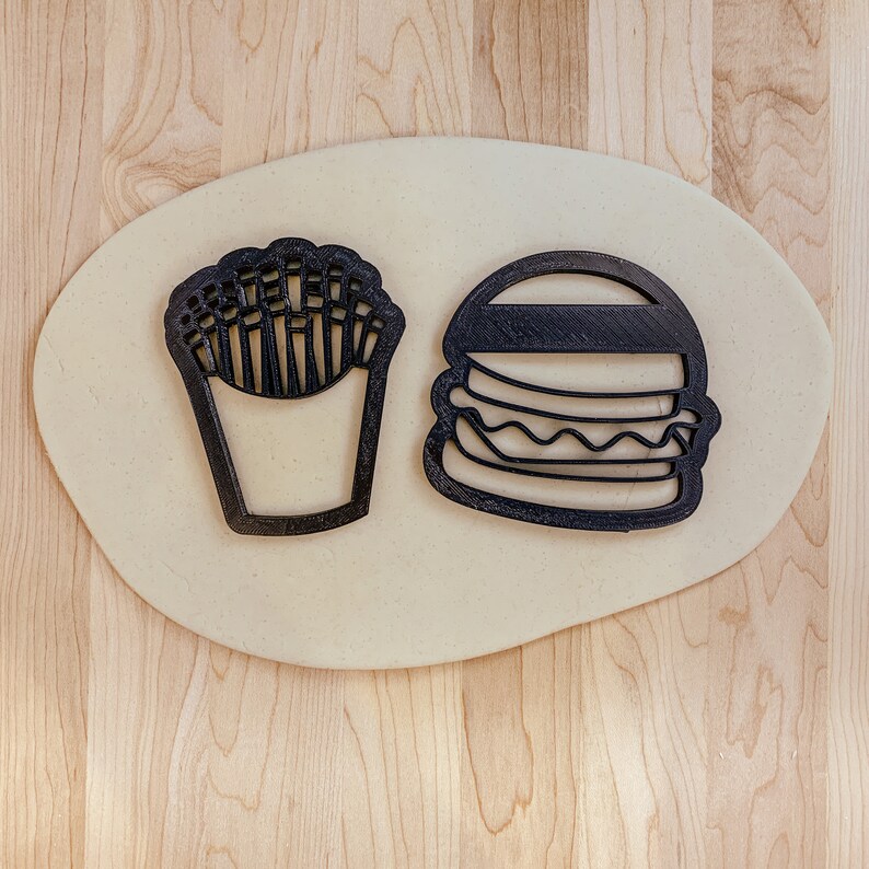 May include: A close-up of two black cookie cutters on a light brown surface. The cookie cutters are shaped like a hamburger and a container of french fries.