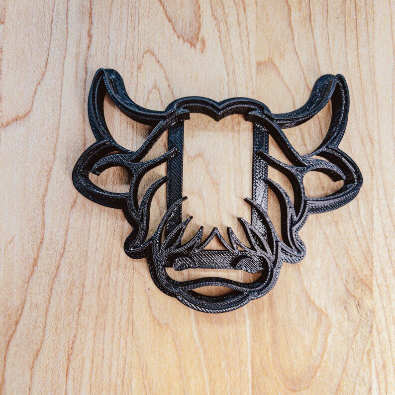 May include: A black, 3D-printed cookie cutter in the shape of a cow's head. The design features detailed horns, floral accents, and a rectangular opening for the cookie. The cutter is placed on a light wood surface.