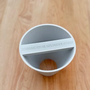 May include: A light grey 3D-printed cylinder form with the text "3DKAMOTION CYLINDER FORM" printed on a horizontal bar across the top. The cylinder is open at both ends and sits on a light brown wooden surface.