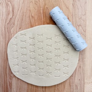 May include: A grey rolling pin with a dragonfly pattern is used to create a pattern on a piece of beige clay.