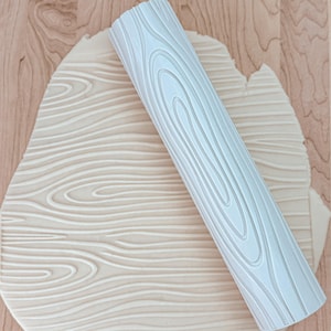 May include: A white silicone rolling pin with a wood grain pattern is shown on top of a piece of fondant that has been rolled with the pin. The fondant has a wood grain pattern.