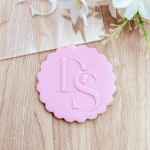 May include: A light pink, scalloped-edge emblem with the embossed letters "DS". The emblem is circular and sits on a light brown wooden surface. White flowers and a clear acrylic piece are in the background.