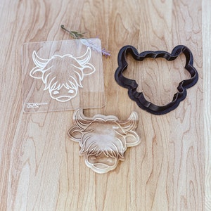 May include: A set of three cookie cutters in the shape of a Highland cow head. The cutters are made of black plastic and are shown on a wooden surface. The cutters are for making cookies or fondant.