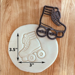 May include: A black plastic roller skate cookie cutter with a white outline of a roller skate on a white surface. The cookie cutter is 7.6 cm wide and 8.9 cm tall.