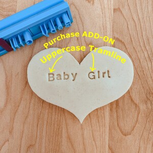 May include: A heart-shaped cookie dough with the words "Baby Girl" stamped into it. The words are in uppercase letters. The dough is on a wooden surface.
