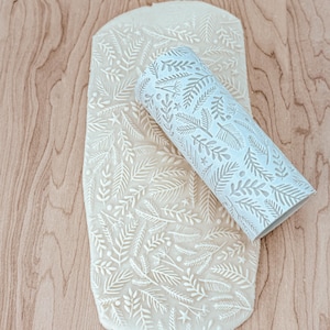 May include: A white rolling pin with a repeating pattern of leaves and sprigs is rolling over a sheet of white dough. The dough is imprinted with the same pattern as the rolling pin.