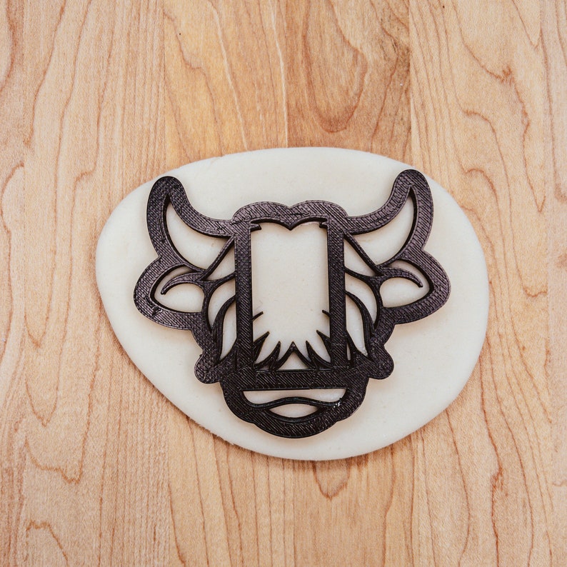 May include: A black, openwork cookie cutter in the shape of a bull's head, resting on a white oval of dough. The cutter's design includes horns, ears, and a central rectangular opening. The background is a light-colored wooden surface.