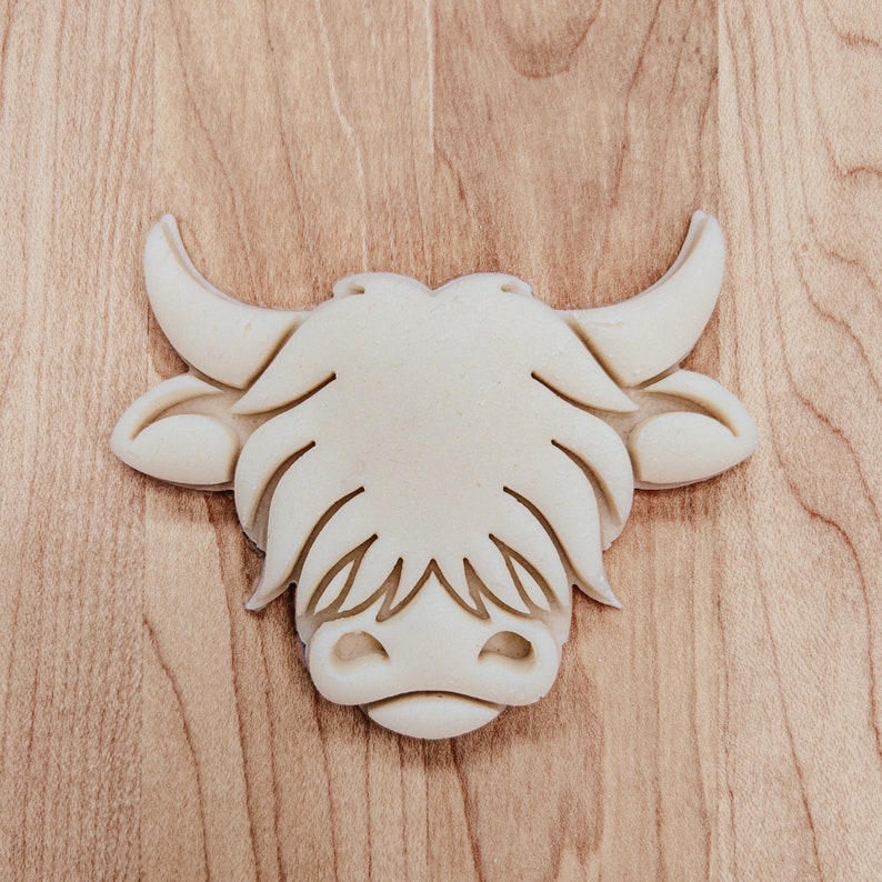 May include: A light-colored, Highland cow head-shaped cookie cutter. The cutter features detailed facial features, including horns, ears, and a fluffy face. The cutter is made of a light-colored material and is set against a wooden background.