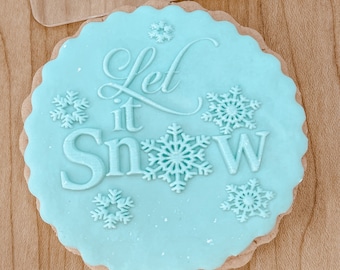 May include: A light blue sugar cookie decorated with white icing and the words "Let it Snow" in cursive. The cookie is decorated with snowflake designs.