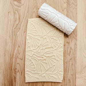 May include: A white rolling pin with a raised leaf pattern design. The rolling pin is on a wooden surface next to a piece of beige clay with the same leaf pattern embossed into it.