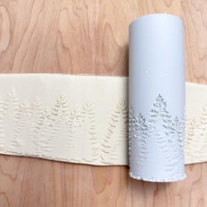May include: A light grey cylindrical clay roller featuring a forest tree design. The roller is used to imprint the tree pattern onto a strip of light-coloured clay. The clay strip is laid on a wooden surface.