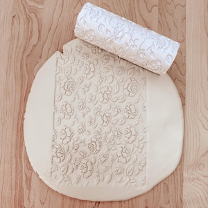 May include: A white, floral-patterned rolling pin lies on a circular piece of dough, showing the embossed rose design. The rolling pin is cylindrical, and the dough is light beige, set against a wooden surface.