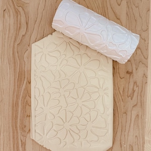 May include: A white, floral-patterned rolling pin and a matching clay slab. The rolling pin is cylindrical, with a raised daisy design. The clay slab has the same daisy pattern pressed into it. The items are on a light wood surface.