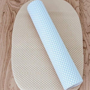 May include: A white silicone rolling pin with a textured pattern of scales. The rolling pin is lying on a beige silicone mat with a similar textured pattern.