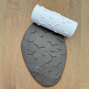 May include: A white rolling pin with a pattern of seagulls is used to create an imprint of seagulls on a gray clay surface.