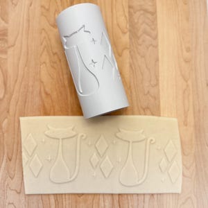 May include: A white rolling pin with a raised design of a cat and stars. The rolling pin is used to create an embossed design on a piece of dough.