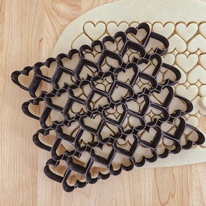 May include: A black heart-shaped cookie cutter with a repeating heart pattern, forming a lattice design. The cutter is pressed into a light-coloured dough, with some cut-out heart shapes visible.