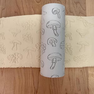 May include: A light grey rolling pin featuring a repeating pattern of mushroom designs. The pin is next to a sheet of dough with matching impressions. The background is a light brown wooden surface. The rolling pin is likely used for baking.