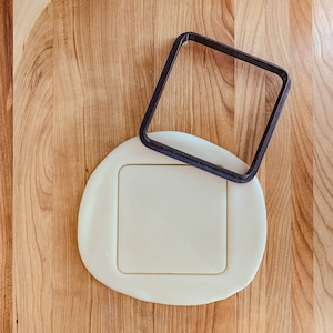 May include: A dark blue square cookie cutter rests on a piece of dough, forming a square shape. The dough is a pale beige colour, and the background is a light brown wooden surface. This image is suitable for baking and crafts.