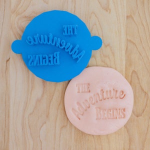 May include: Two stamps, one blue and one pink, with the text "The Adventure Begins" in a cursive font.