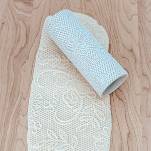 May include: A white rolling pin with a lace pattern embossed on the surface. The rolling pin is laying on a piece of white fondant with a lace pattern.