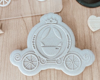 May include: A white, detailed, plastic cookie cutter in the shape of a carriage with a crown on top. The carriage has wheels and a curved design.