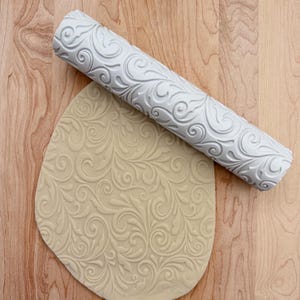 May include: A white embossed rolling pin with a floral design is used to create a pattern on a sheet of dough.