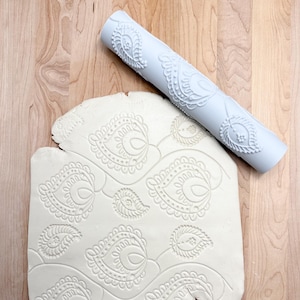 May include: A light blue patterned rolling pin and a piece of clay with an embossed paisley design. The rolling pin is cylindrical and features a repeating paisley pattern. The clay has the same pattern pressed into it.