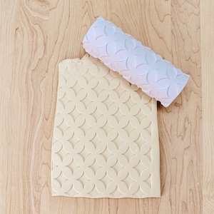 May include: A white rolling pin with a floral pattern rests on a piece of beige dough, which has been imprinted with the same design. The dough is on a light wood surface, creating a contrast between the textures and colors.