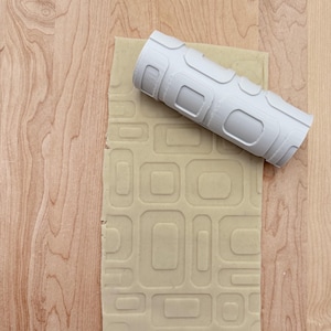 Peut inclure: Un rouleau à pâtisserie en plastique blanc avec un motif géométrique roule sur une feuille de pâte beige. La pâte a été embossée avec le même motif géométrique.