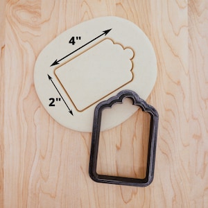 May include: A 4 inch by 2 inch cookie cutter in the shape of a tag with a decorative edge. The cutter is made of metal and is shown on a wooden surface.