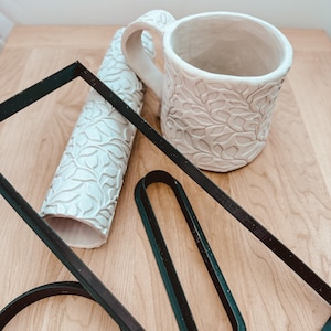 May include: A white ceramic mug with a leaf pattern, a white ceramic rolling pin with a leaf pattern, and two black plastic stencils. The stencils are shaped like a rectangle and an oval.
