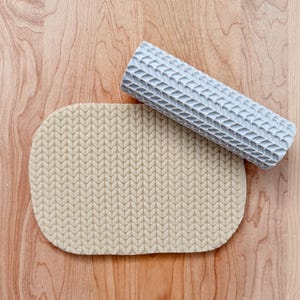 May include: A light grey rolling pin with a textured knit pattern is positioned beside a piece of dough with the same pattern. The dough is a light beige colour. The rolling pin is made of a hard material, and the dough is soft.