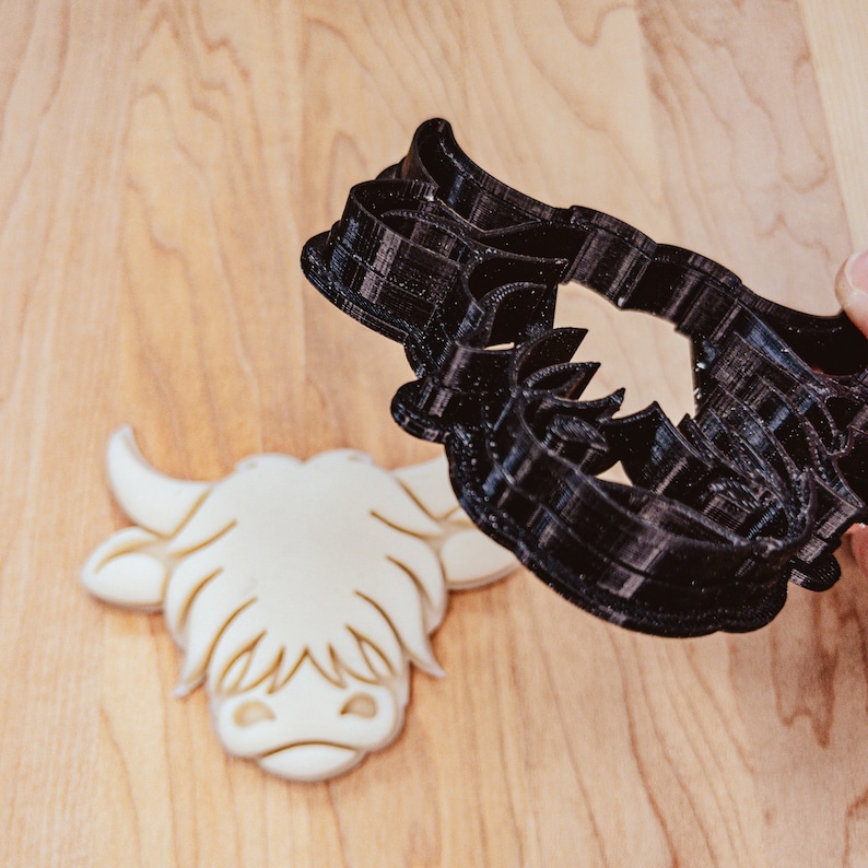 May include: A black plastic Highland cow cookie cutter next to a cream-colored cookie in the shape of a Highland cow head. The cookie cutter is held by a hand. The background is a light brown wooden surface.