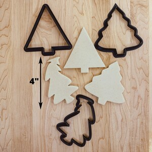 May include: Five cookie cutters in the shape of Christmas trees. The cutters are made of black metal and are 10 cm tall. The cutters are on a wooden surface.