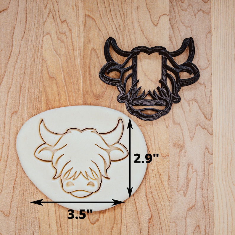 May include: A black Highland cow cookie cutter next to a cookie with the same design. The cookie cutter is 3.5 inches wide and 2.9 inches tall. The cookie is in the shape of a Highland cow's head.