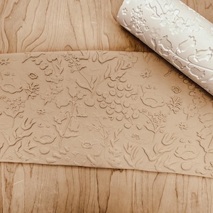 May include: A beige floral embossed rolling pin and a sheet of embossed paper with a similar floral design. The rolling pin is rolled up, and the paper is laid flat, showing a detailed pattern of flowers and leaves.